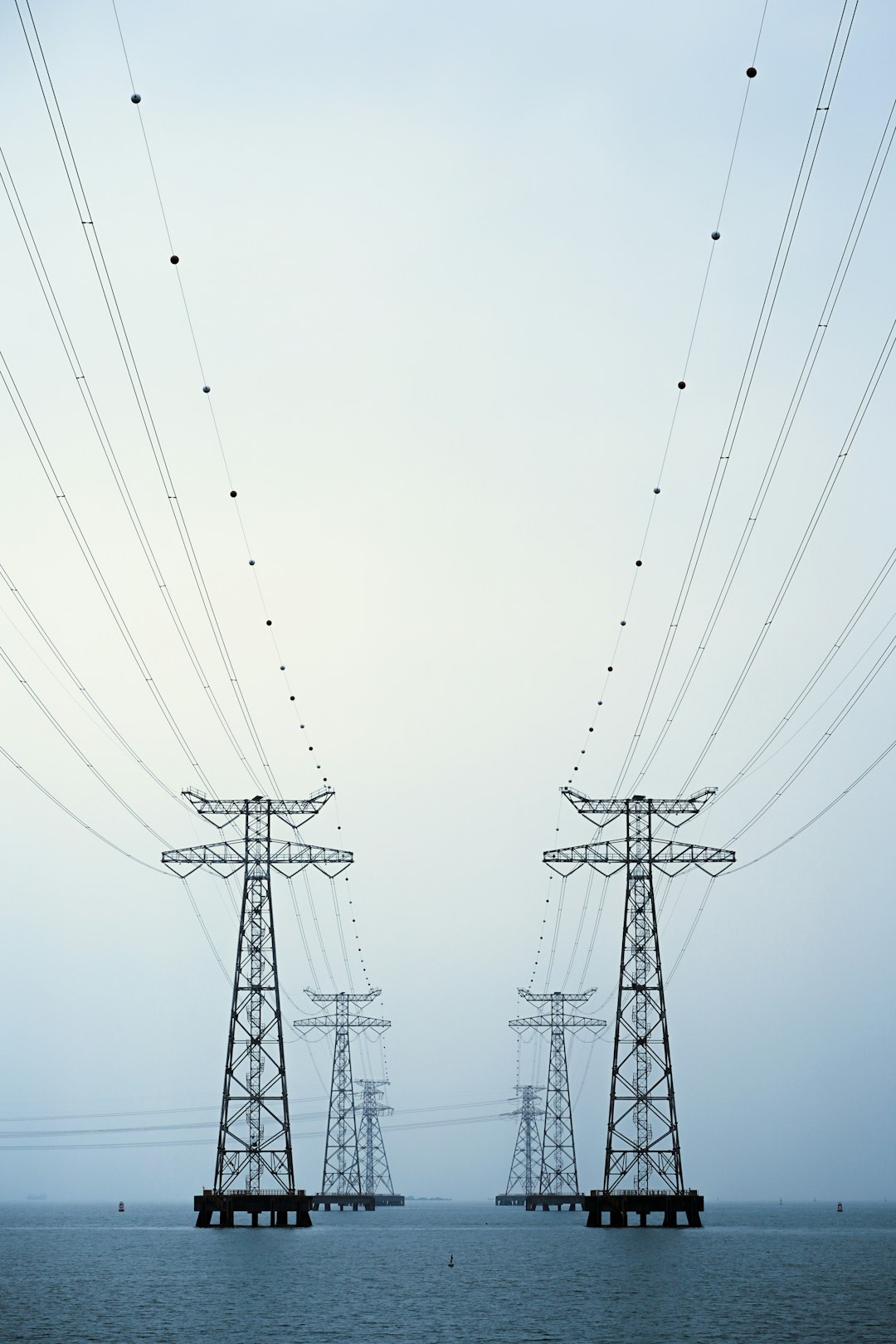 Two lattice electricity pylons carry a cross-sea power line over the calm waters of Shenzhen Bay, Guangdong, China. Soft morning haze flattens the colour palette, turning the steel towers and sweeping cables into a minimalist industrial seascape with strong leading lines.